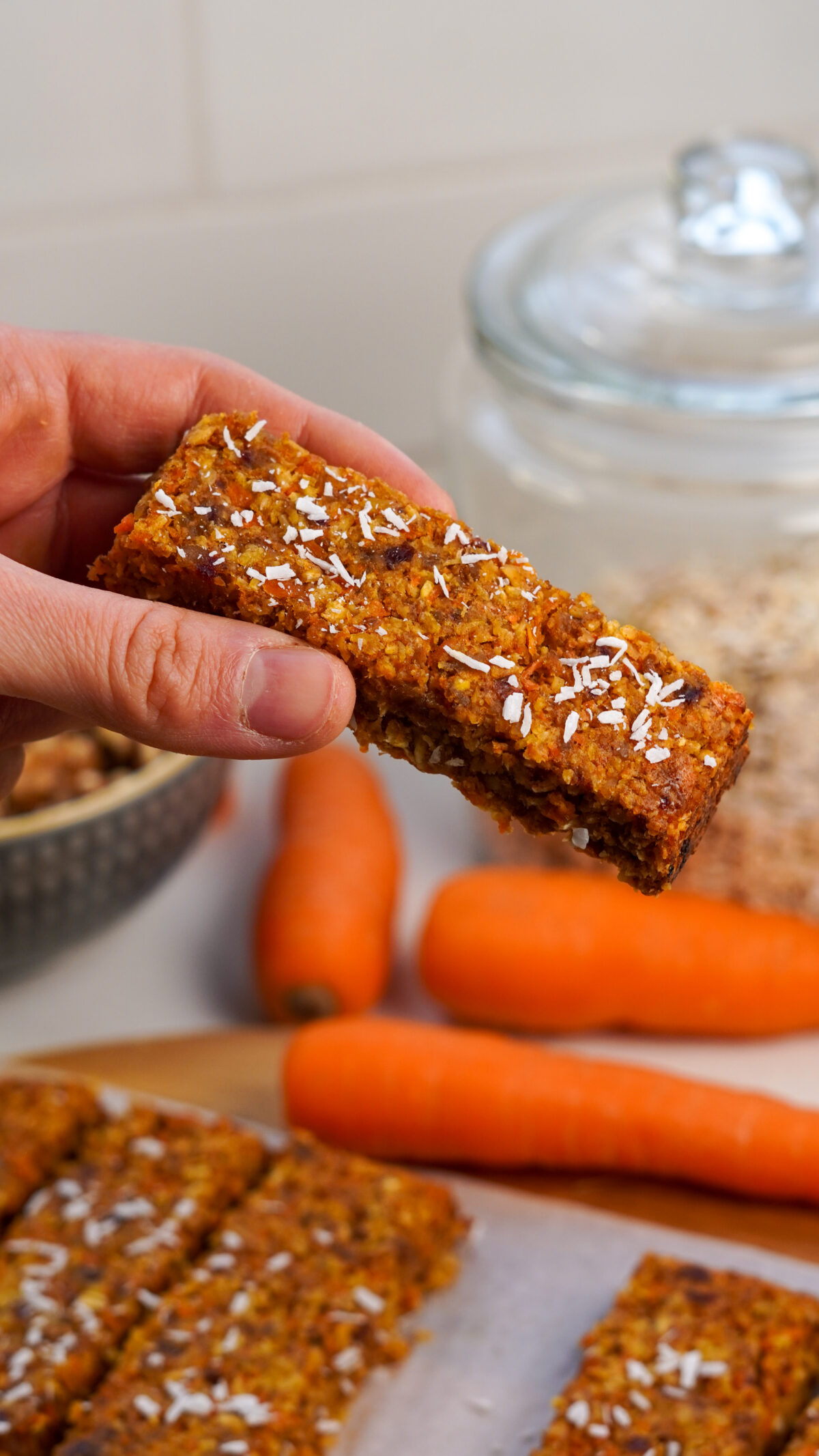 Hand holding a healthy carrot cake bar with three more bars and carrots in the background