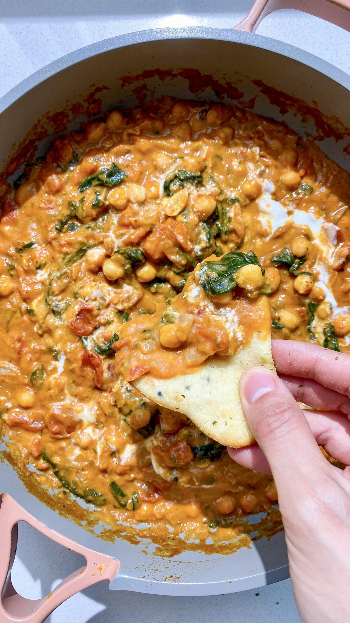 Overhead shot of a creamy chickpea curry in a pan, with one hand holding a small piece of naan topped with curry. A warm, cosy and inviting scene showcasing this easy one-pan meal.
