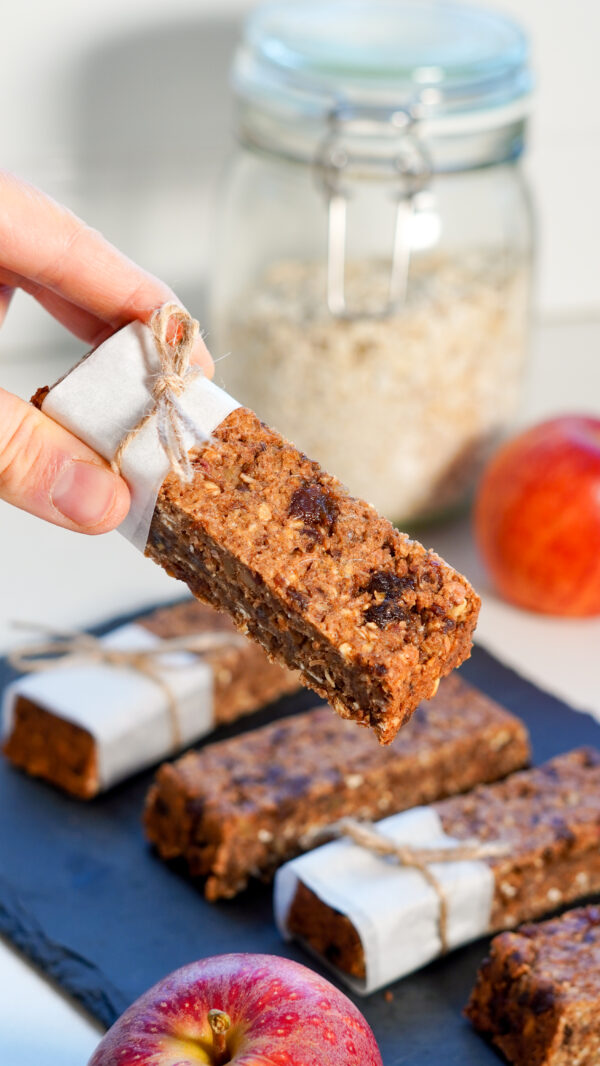 Two fingers holding one apple oat bar with more bars, a jar of oats, and a red apple in soft focus background