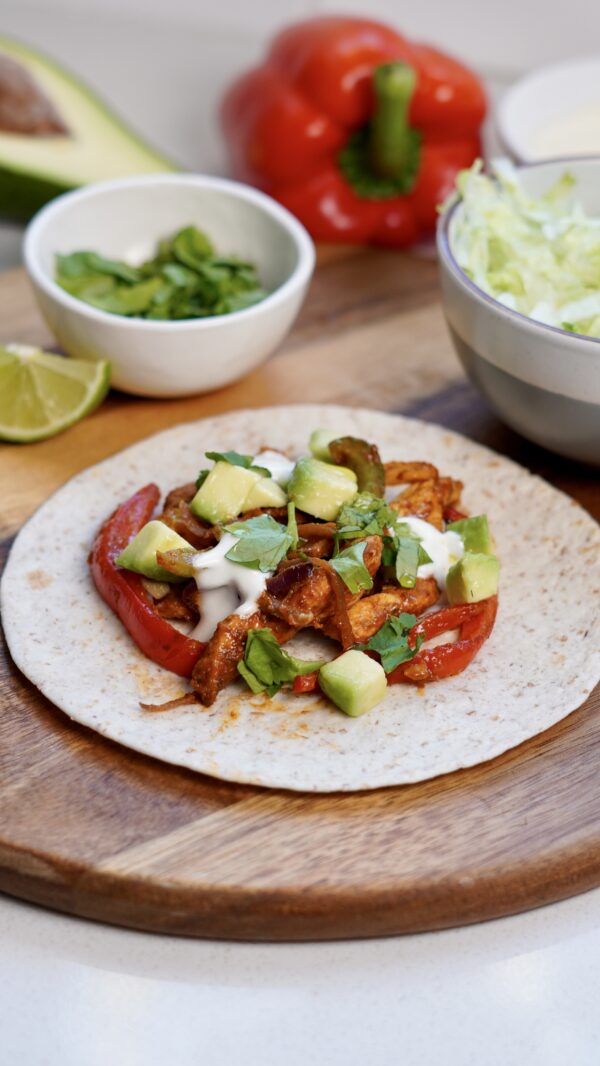 Open air fryer fajita with chicken, peppers, and onions on a chopping board, surrounded by fresh ingredients in the background