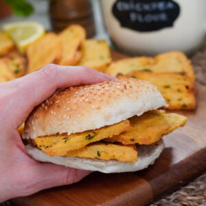 Hand holding a Sicilian panelle sandwich in a burger bun, with more panelle in soft focus in the background.