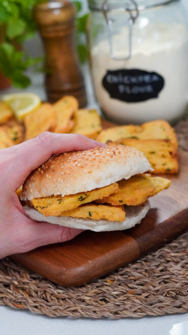 Hand holding a Sicilian panelle sandwich in a burger bun, with more panelle in soft focus in the background.
